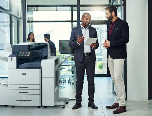 Dos hombres en una oficina conversan sobre algo en una tableta junto a una impresora multifunción en color Xerox® AltaLink® C8200 Series